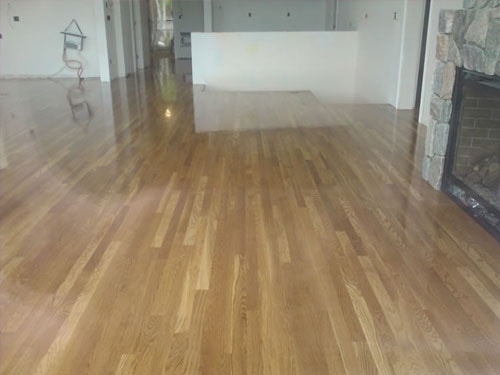 Freshly finished warm oak hardwood floor with high-gloss sheen in open living area with stone fireplace