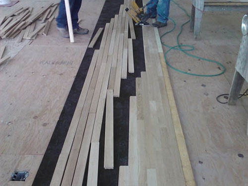 Worker nailing hardwood flooring planks over black felt underlayment during installation
