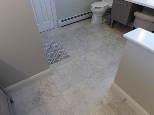 Large-format light gray marble-look tile floor in bathroom with gray vanity and diamond-pattern rug