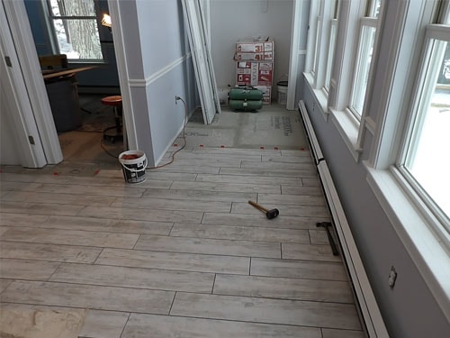 Wood-look porcelain tile installation in progress in sunroom hallway with grout bucket and rubber mallet