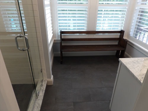 Charcoal slate tile bathroom floor with wood bench, glass shower enclosure, and plantation shutters