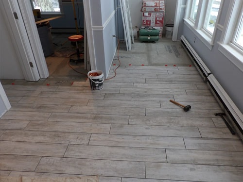 Light gray wood-look tile installation in progress in sunroom with grout bucket, rubber mallet, and baseboard heaters