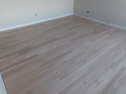 Large unfinished white oak hardwood floor in empty bedroom with multiple windows and white walls