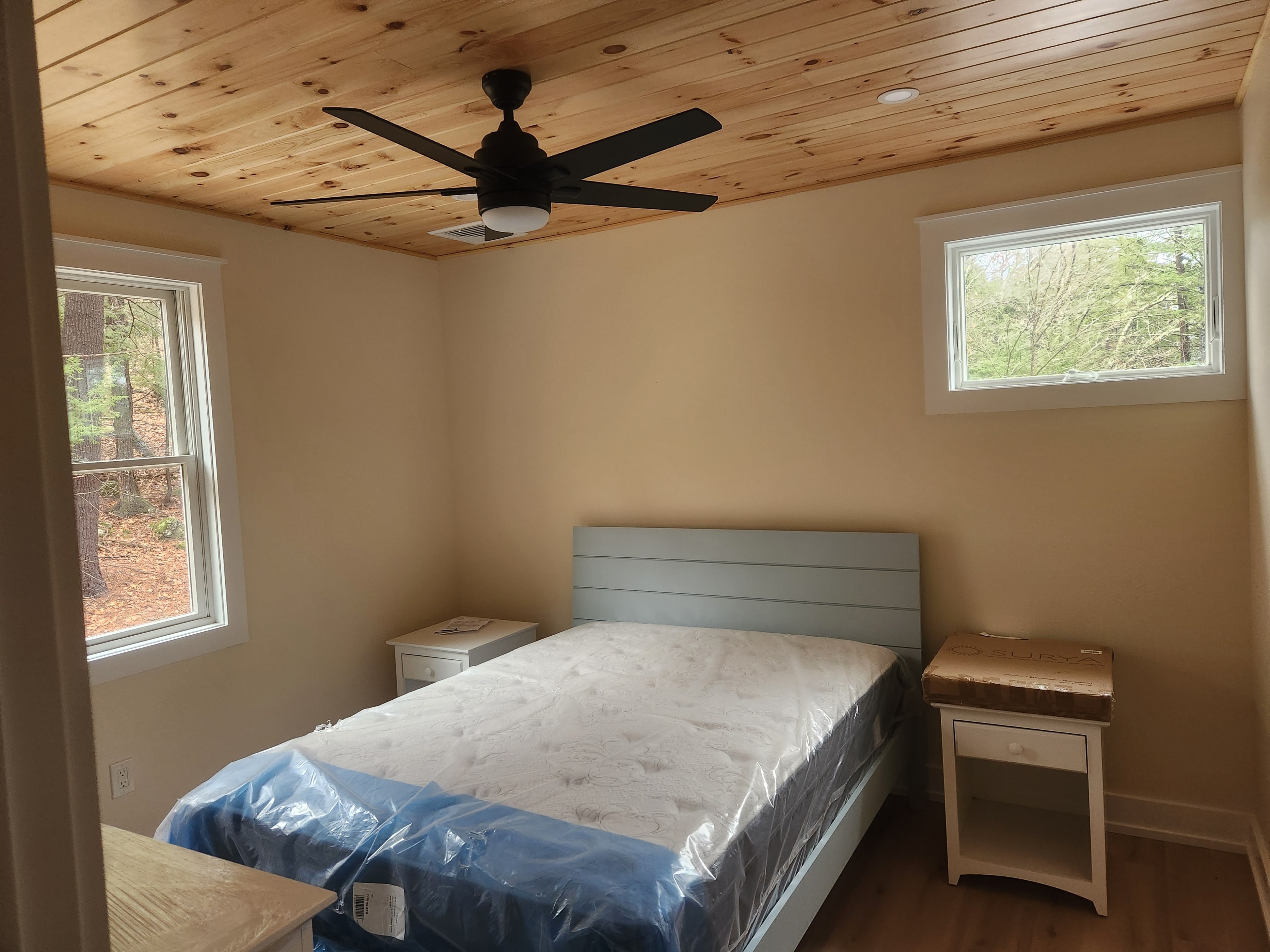 Finished bedroom with knotty pine ceiling, blue-gray headboard, and wooded views through double windows