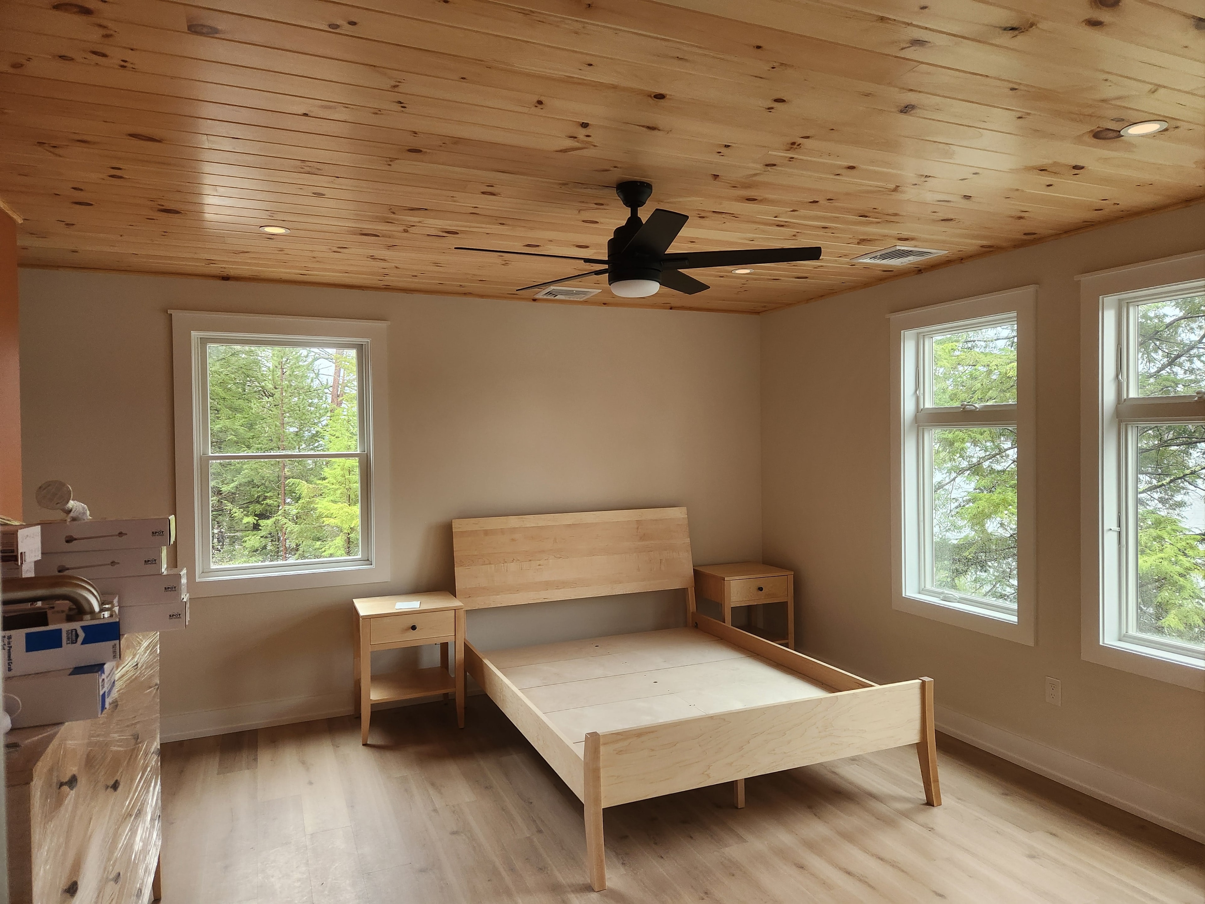 Finished bedroom with knotty pine ceiling, maple bed frame, and wooded views through large windows
