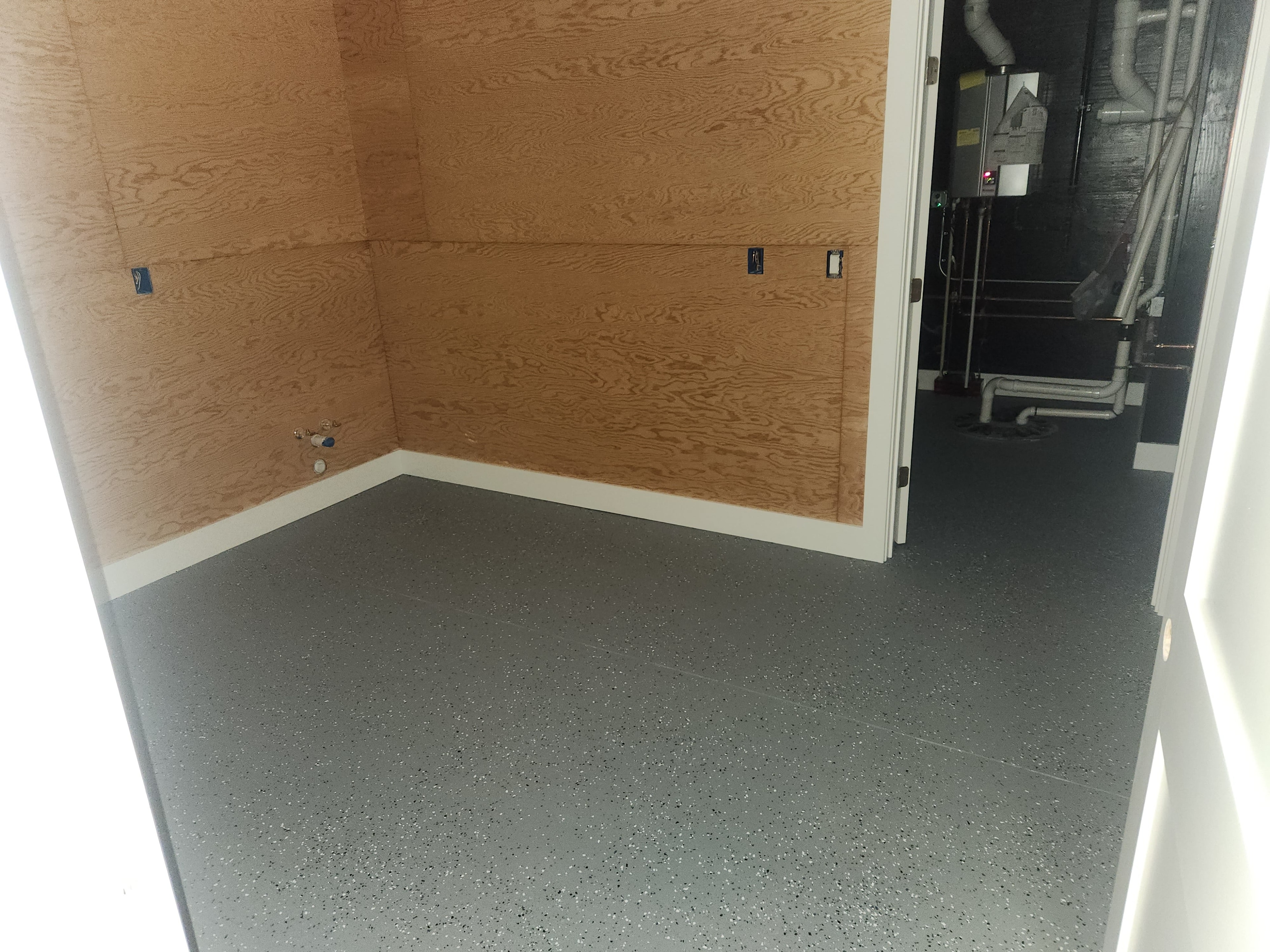 Second angle of workshop room showing epoxy chip floor, OSB walls, and mechanical room beyond doorway