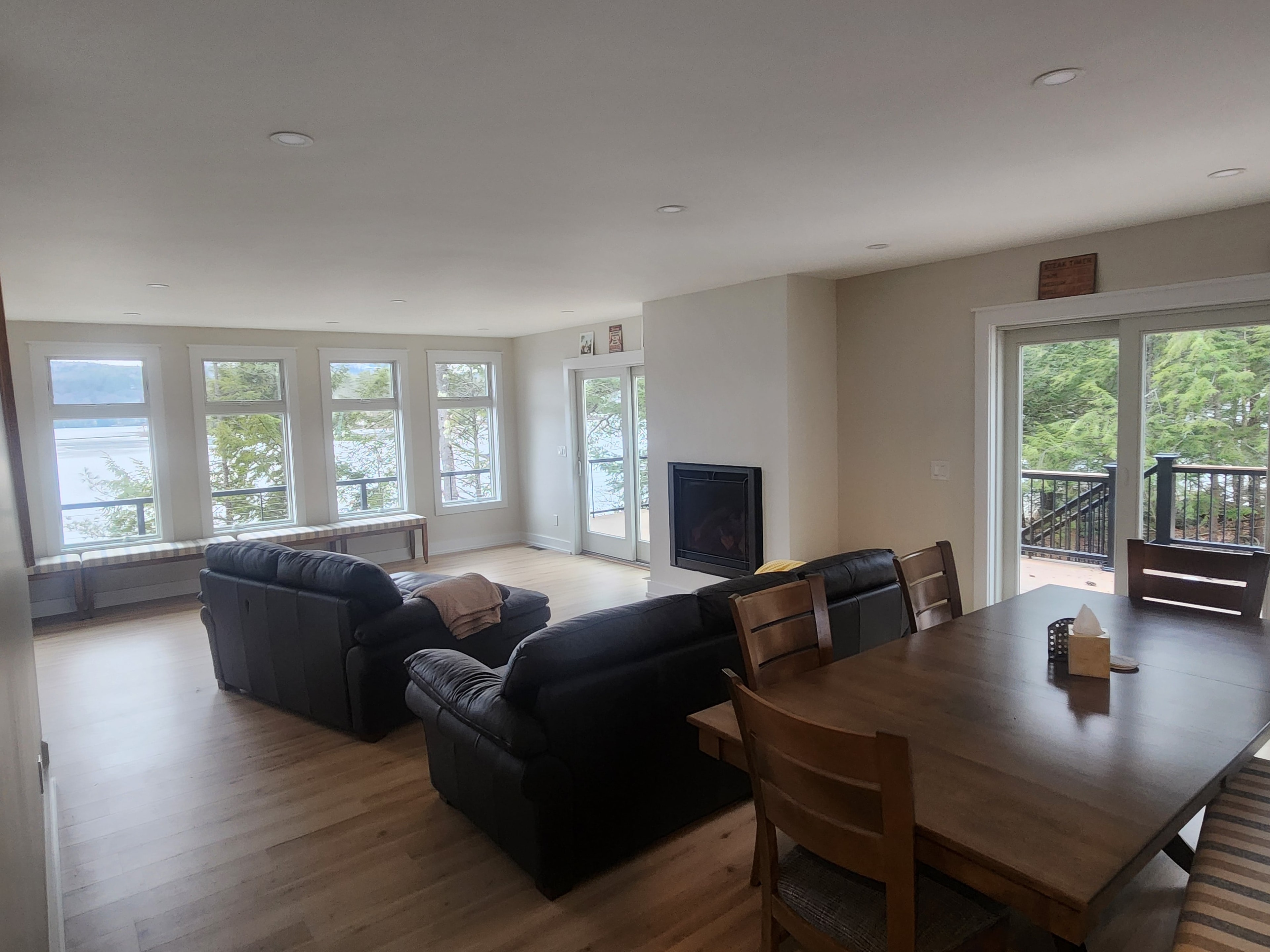 Finished open-plan lakeside living and dining room with hardwood floor, gas fireplace, and panoramic lake and forest views