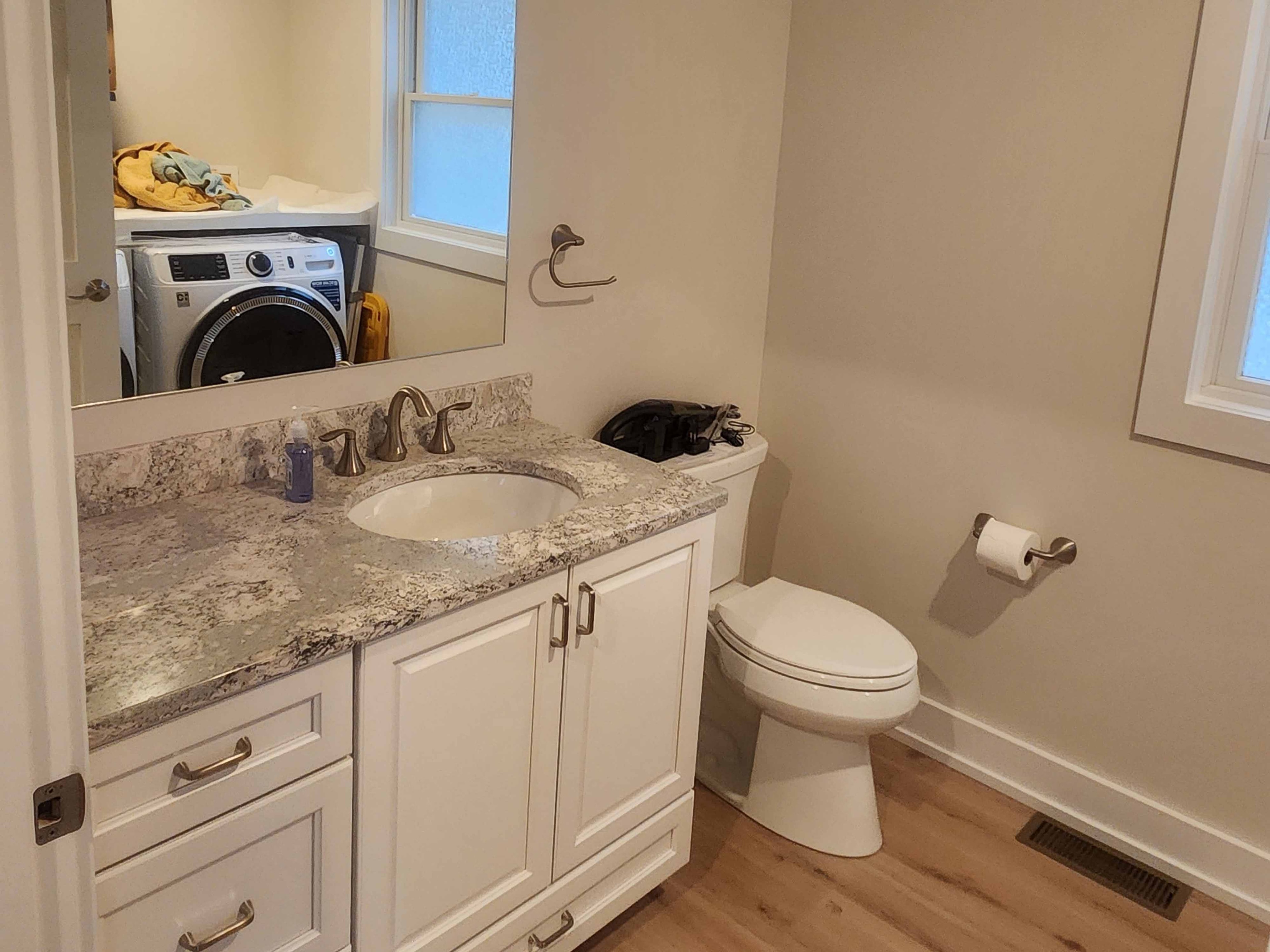 Finished bathroom with white shaker vanity, granite countertop, brushed nickel fixtures, and LVP flooring adjacent to laundry room