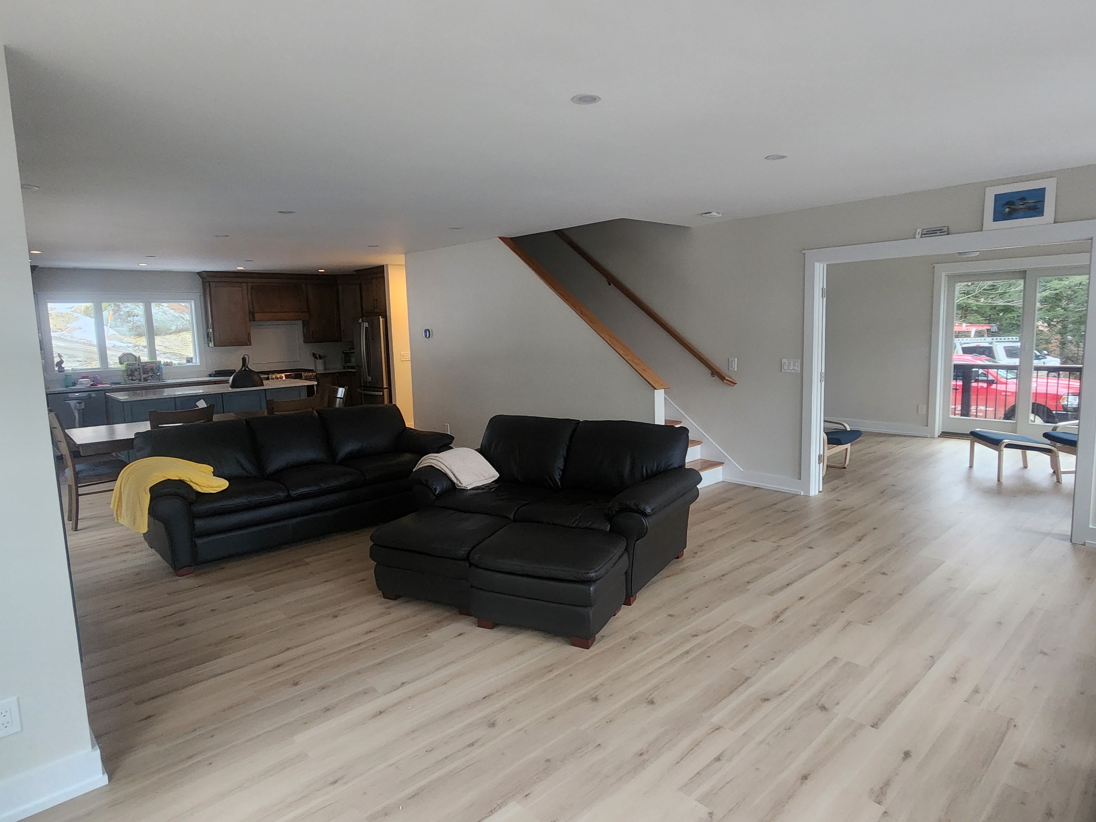 Finished open-plan living room with LVP flooring, staircase with wood handrail, kitchen beyond, and sliding door to deck