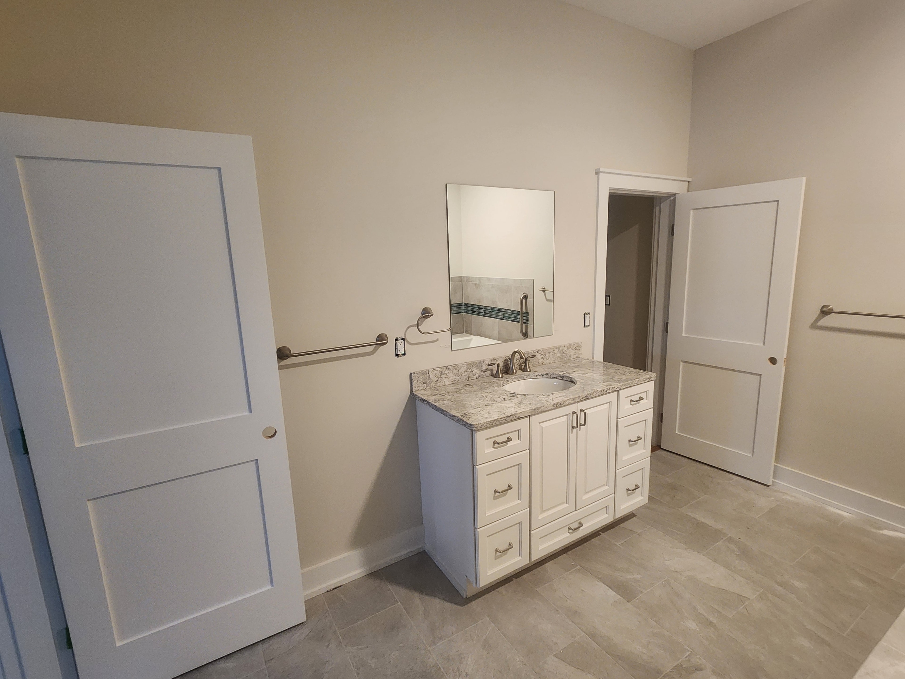 Spacious new construction bathroom with white shaker vanity, light granite countertop, large-format tile floor, and two white panel doors