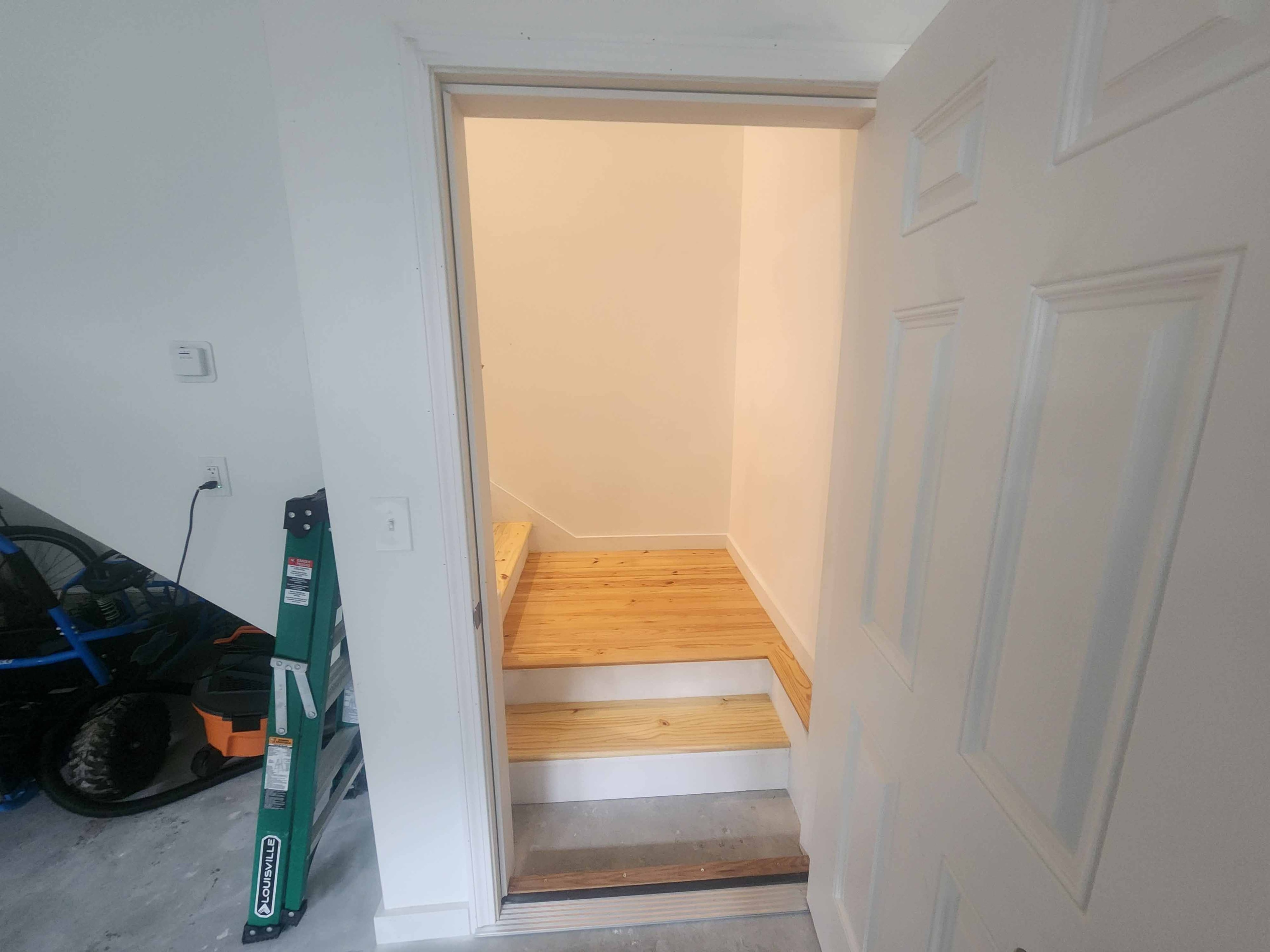 New construction interior staircase with pine treads viewed from garage entry with green ladder and white panel door