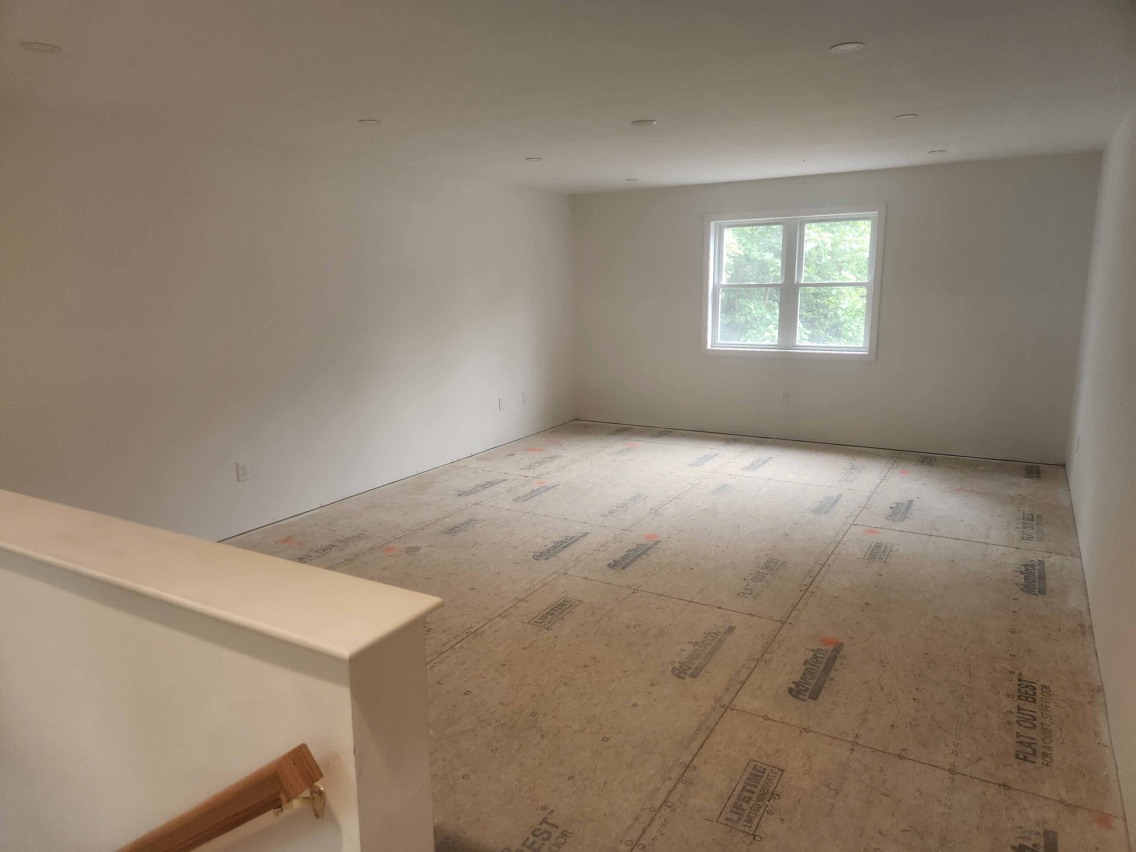 New construction bedroom with white walls, recessed lighting, wooded window view, and Advantech subfloor with stair railing in foreground