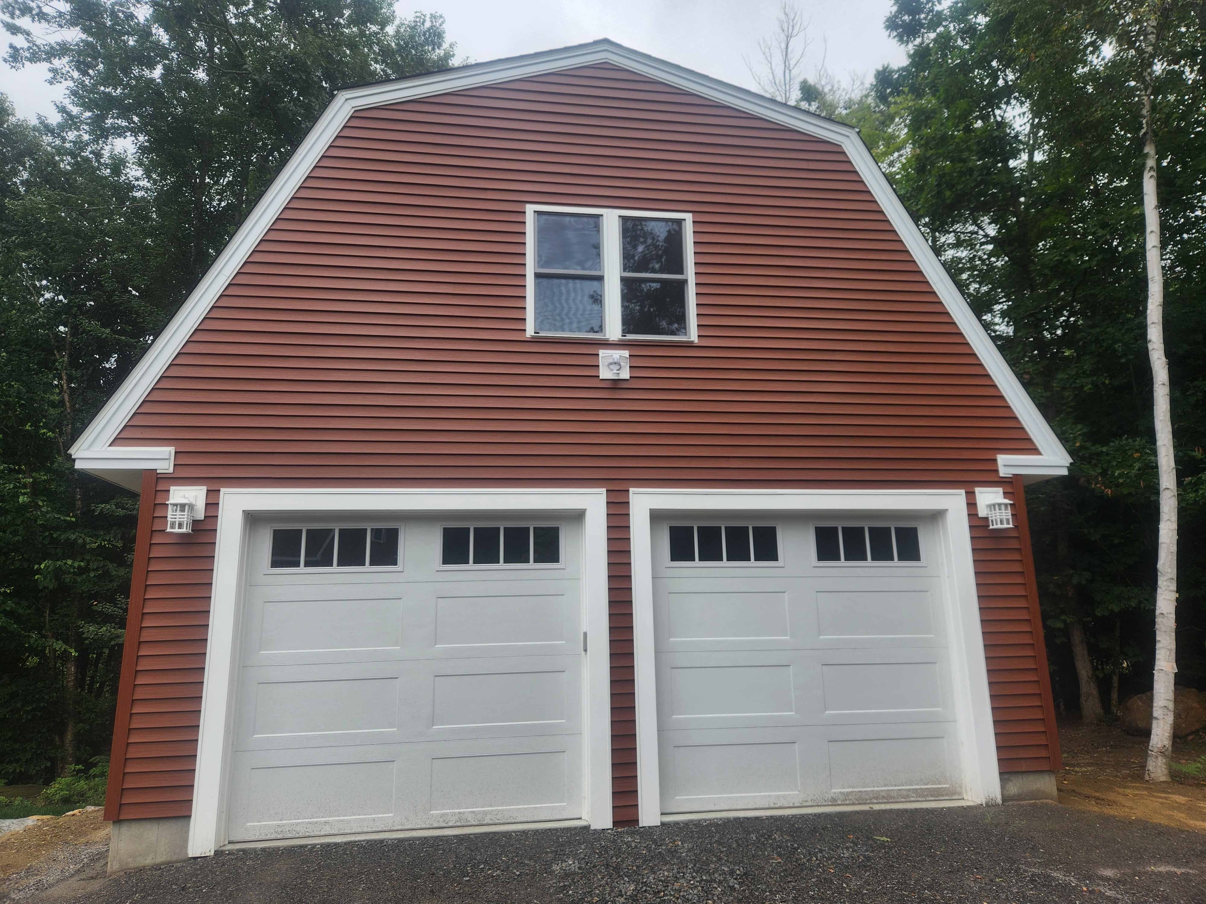 Newly completed gambrel-style two-car garage with red vinyl siding, white garage doors, and upper loft window surrounded by birch trees