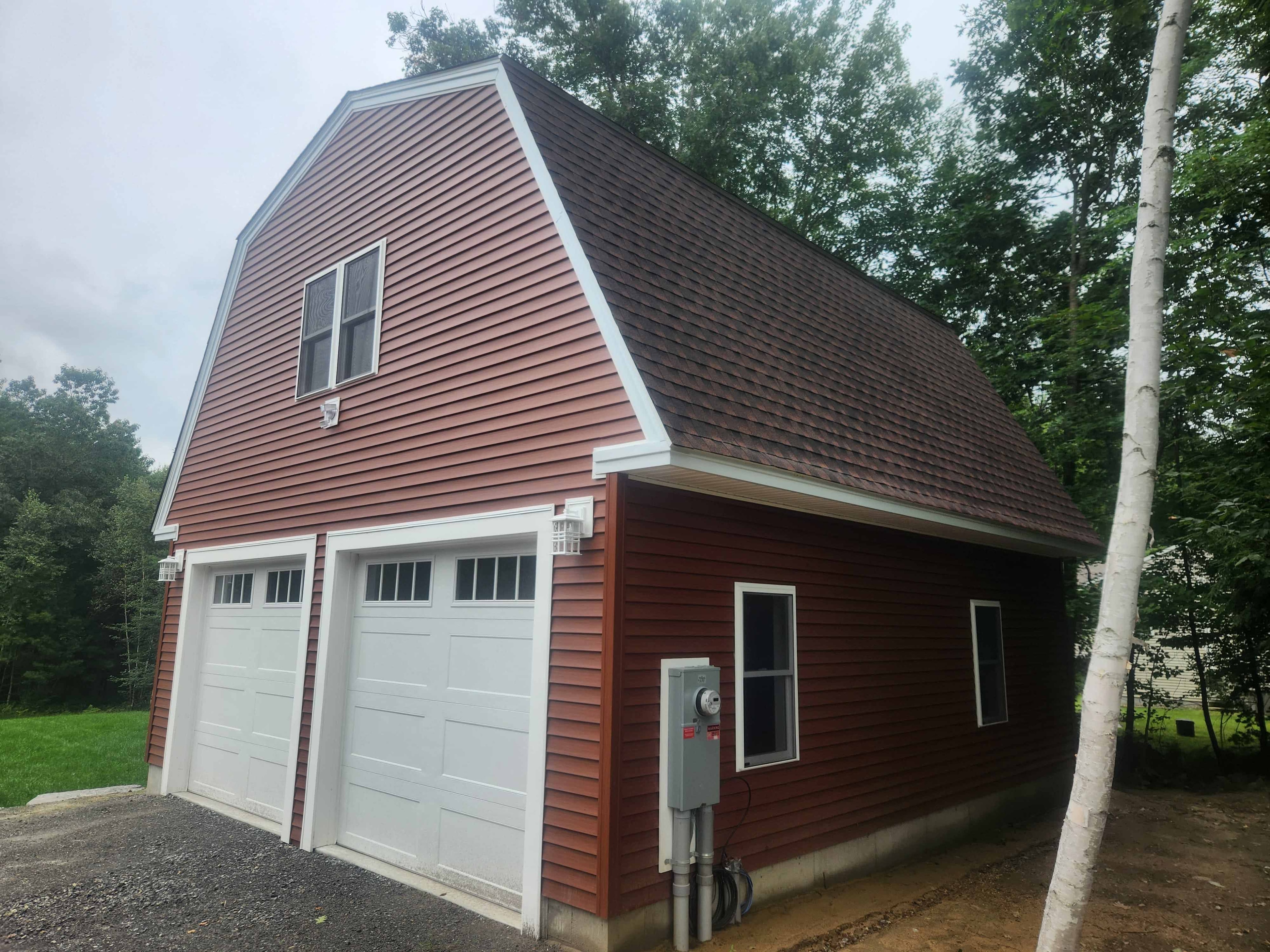Three-quarter exterior view of completed gambrel garage showing red vinyl siding, brown architectural shingles, electrical service, and side windows
